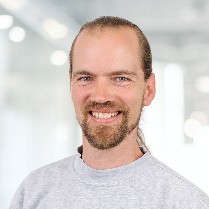 Porträt eines lächelnden Mannes mit Bart vor unscharfem Hintergrund.