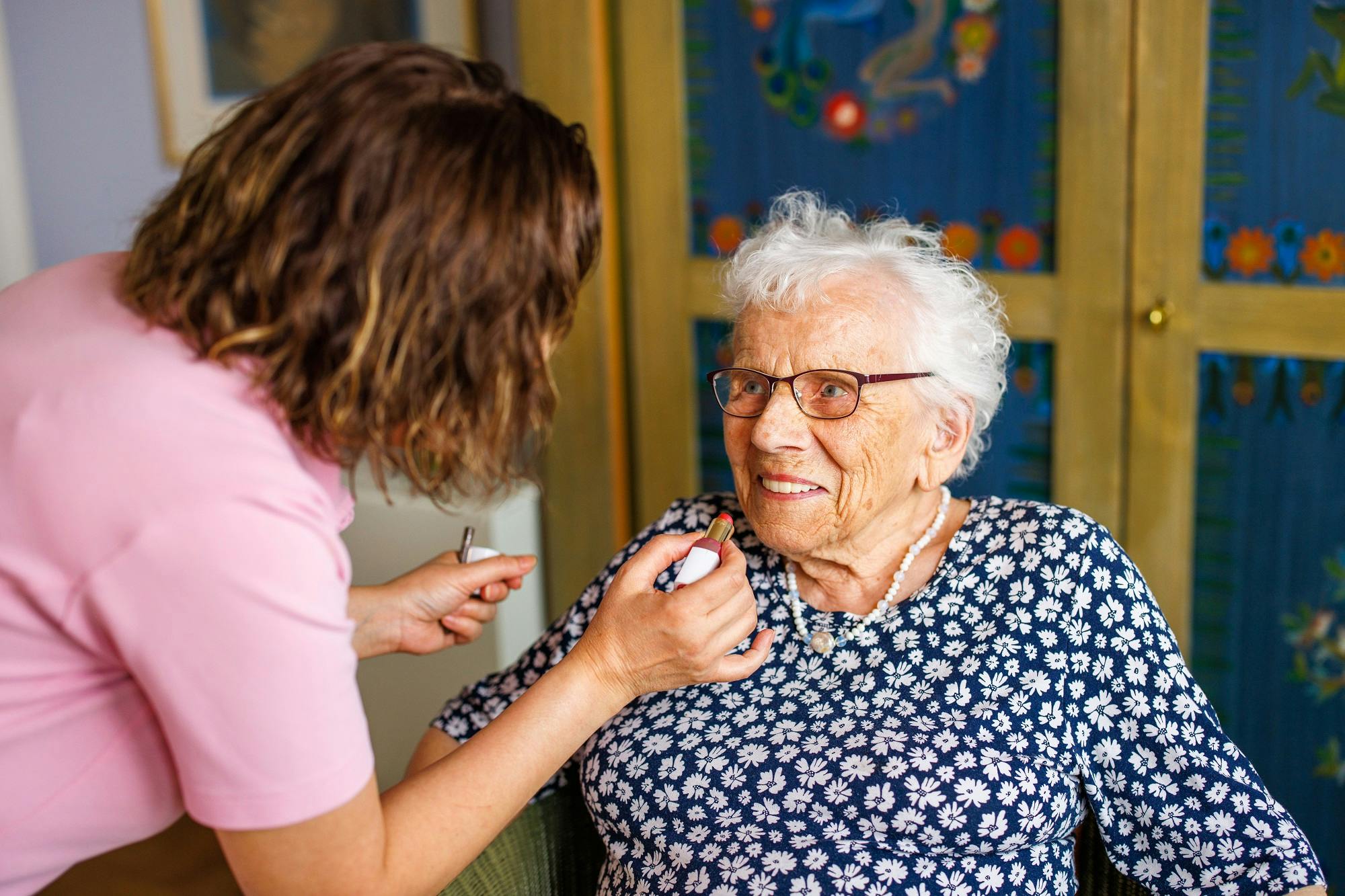 Eine ältere Dame mit Brille wird von einer Pflegekraft mit einem Lächeln im Gesicht versorgt.