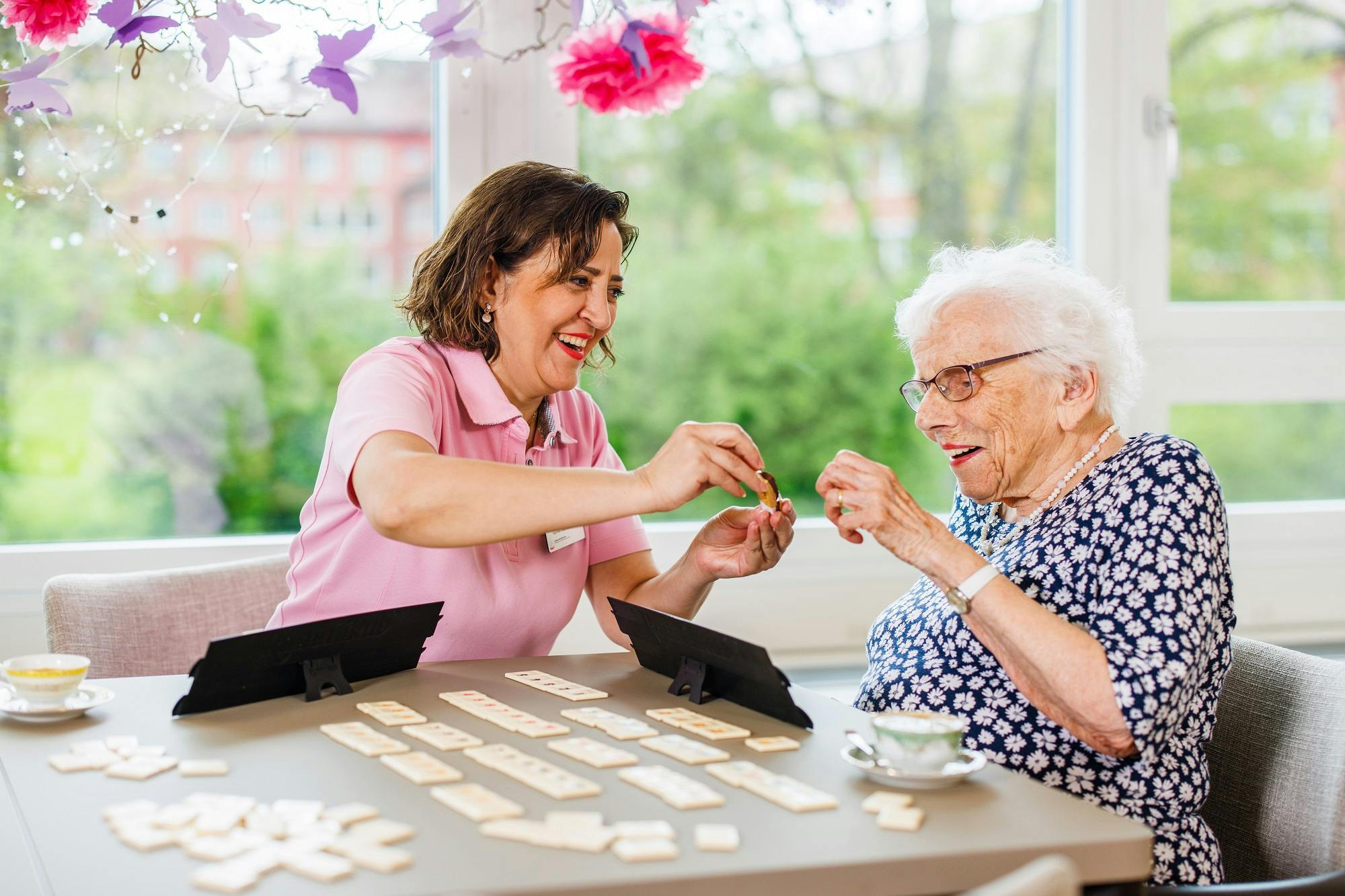 Pflegekraft spielt lachend mit Seniorin Domino in einem hellen Raum.