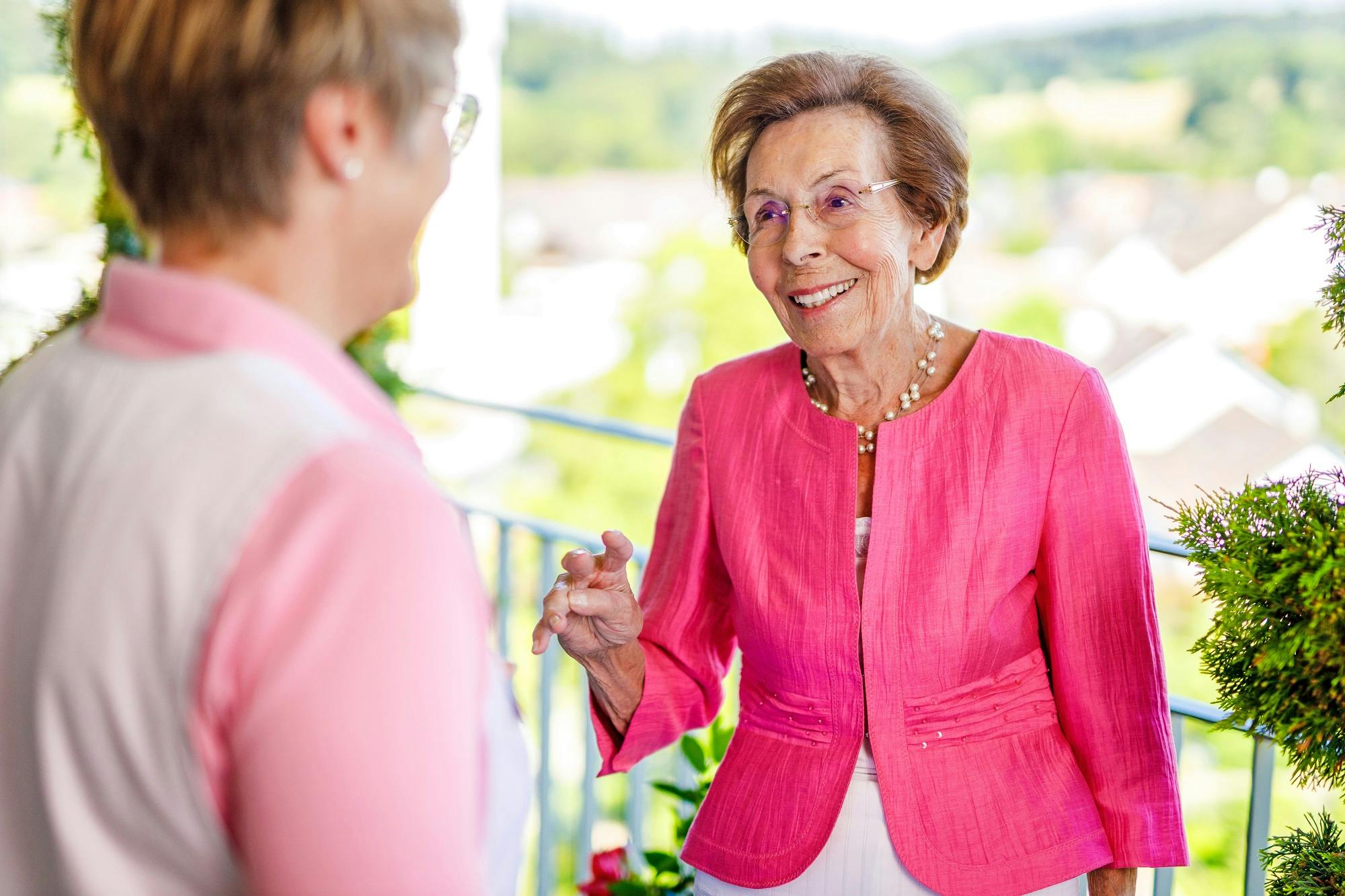 Alt-Text: Ältere Dame in rosa Jacke unterhält sich lächelnd mit einer anderen Person im Freien.