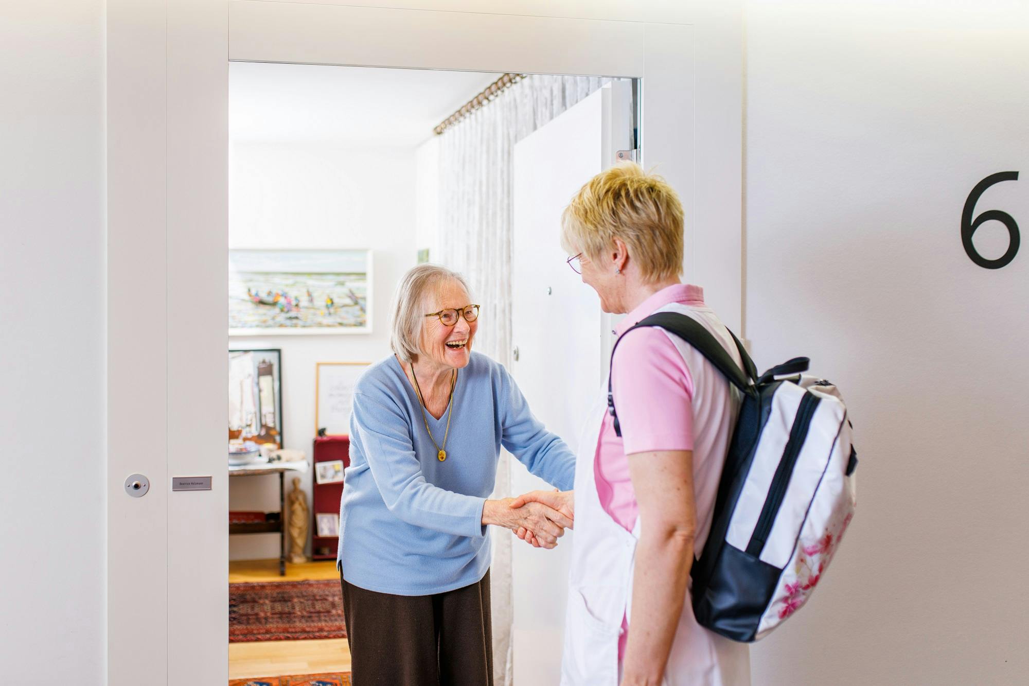 Zwei ältere Damen begrüßen sich lächelnd mit einem Handschlag an einer Haustür.